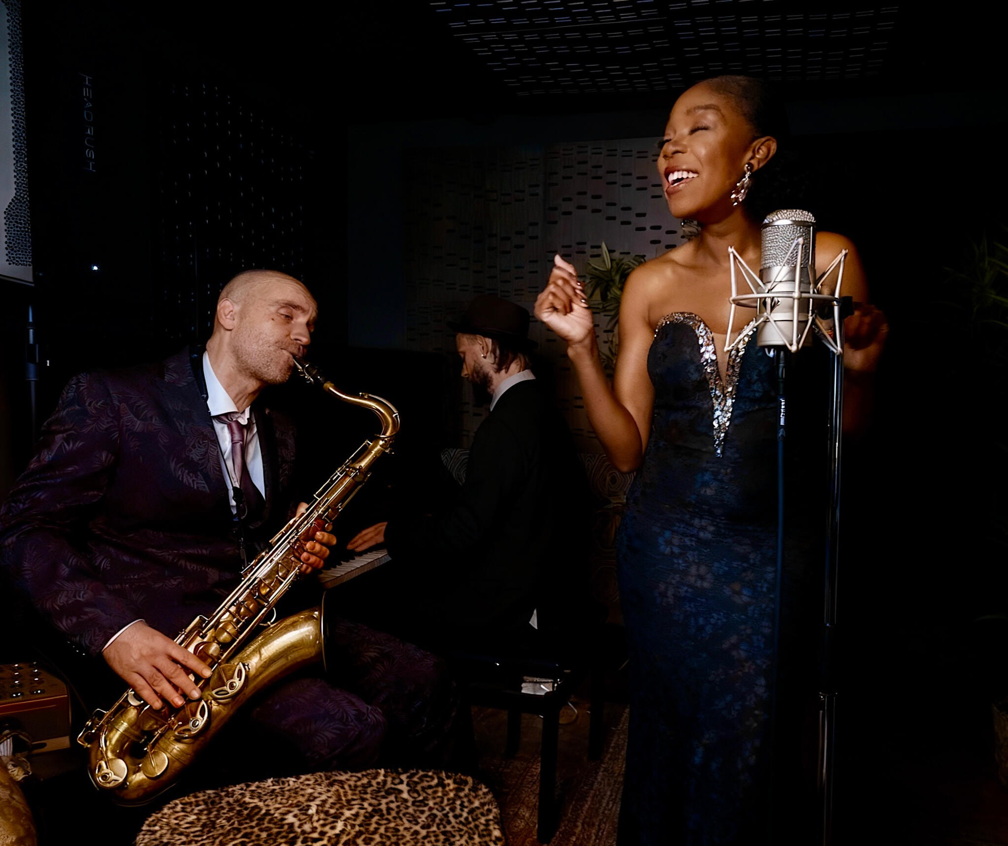 A vocalist sings into a studio microphone while a saxophonist plays on the left and a pianist performs in the background. The scene shows the trio in a warm, low-lit setting that reflects their long collaboration and intimate musical style.