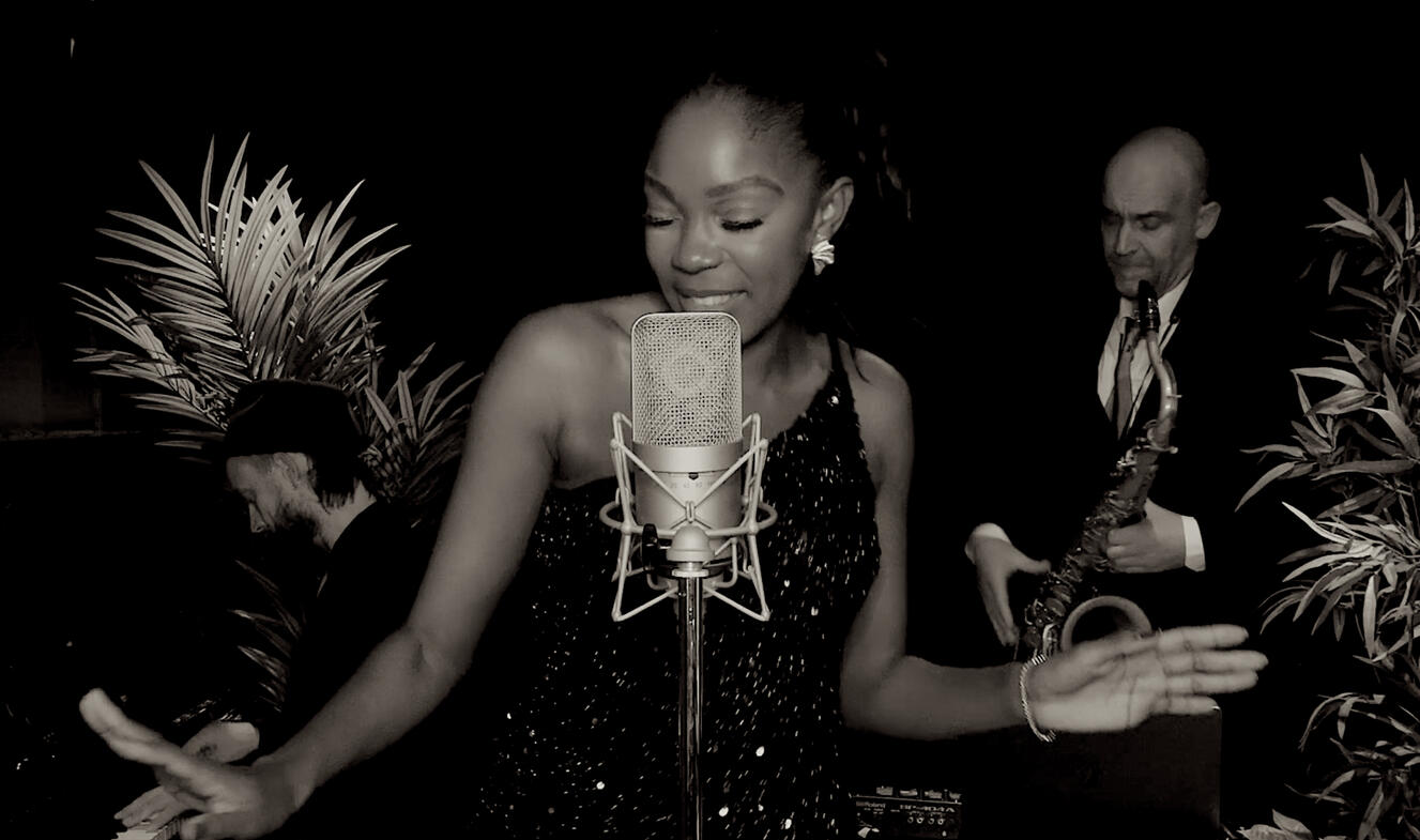 Black and white photo of a vocalist singing into a vintage-style microphone with a pianist on the left and a saxophonist on the right. The trio performs in a dark studio surrounded by plants, creating a refined and atmospheric mood for the homepage.